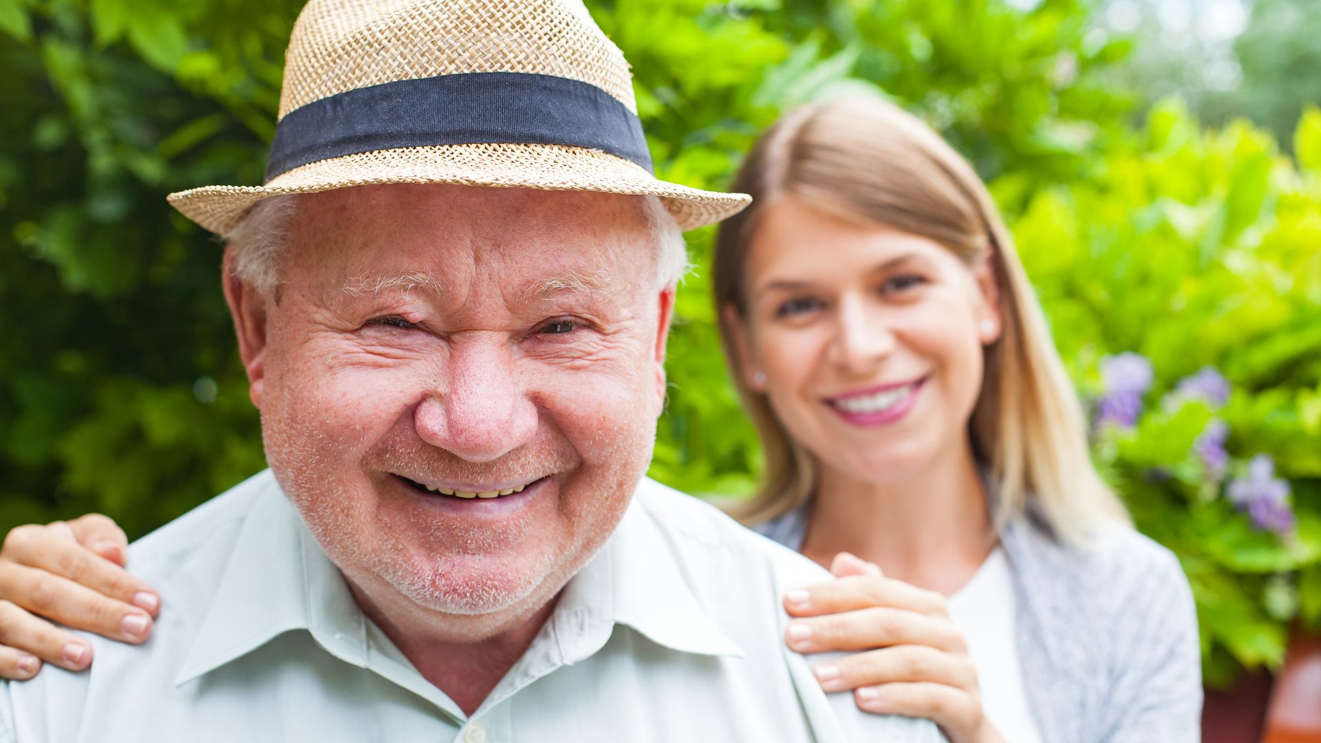 Starszy mężczyzna w kapeluszu i młoda kobieta z uśmiechem stoją razem w ogrodzie. Ciepła relacja, wspólna chwila.