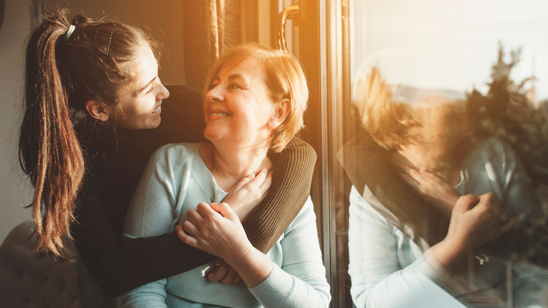 Mama i córka w czułym uścisku, siedzą razem przy oknie w ciepłym świetle zachodzącego słońca.